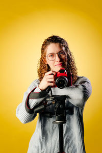 Portrait of young woman holding camera against yellow background