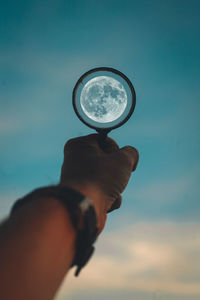 Cropped hand holding magnifying glass against sky