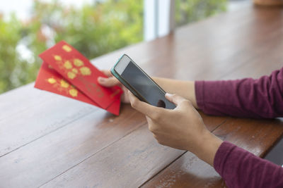 Cropped hands of person using mobile phone while holding greeting cards on table