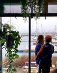Rear view of mother and young son standing by glass window looking at harbour
