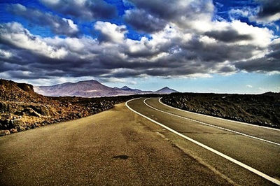 Empty road by mountains against cloudy sky