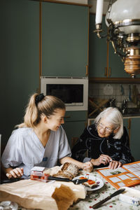 Smiling nurse teaching senior woman at dinning table in kitchen