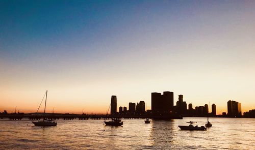 Silhouette sailboats in sea by buildings against clear sky during sunset