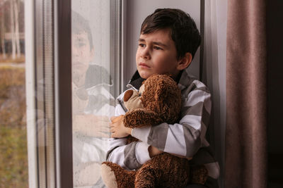 Sad boy looks out the window hugging a teddy bear. bad weather mood, depression