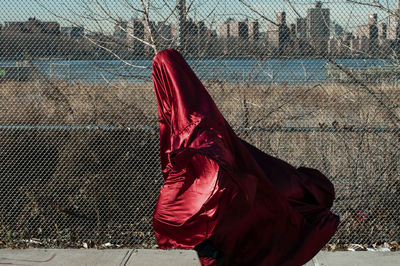 Person wrapped in red fabric while walking on footpath by chainlink fence in city