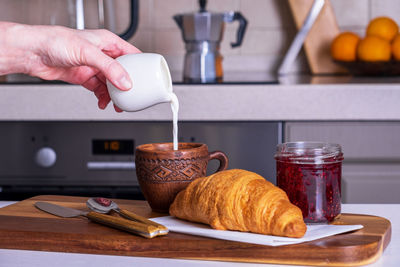 Served morning breakfast with croissant, jam and cup of coffee pouring cream in woman's hand