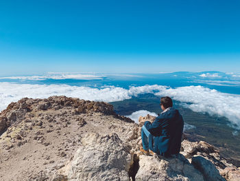 Rear view of man sitting on rocks against blue sky