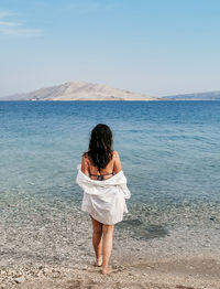 Rear view lifestyle image of stylish young woman in white shirt standing on beach and looking at sea