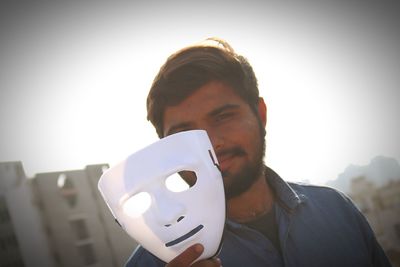 Portrait of young man holding mask against sky