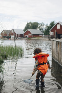 Rear view of boy wearing life jacket while standing in lake with fishing rod during summer