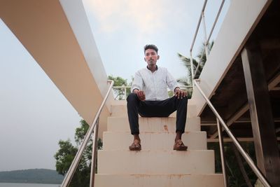 Low angle view portrait of young man sitting on staircase against sky