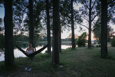 Young woman sitting on hammock amidst trees by lake