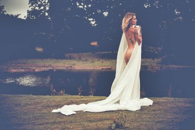 Naked woman holding white fabric while standing at lakeshore