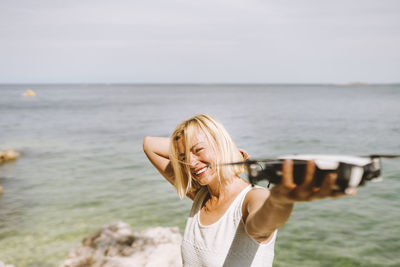 Smiling woman holding drone while standing against sea