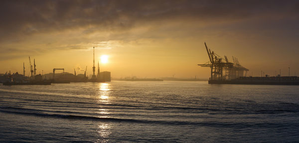 Cranes at commercial dock against sky during sunset