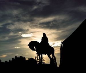 Silhouette man riding horse in city against sky during sunset