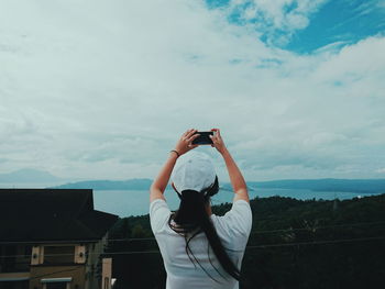 Rear view of woman photographing lake through smart phone against cloudy sky