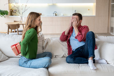 Cheerful girlfriend sitting on sofa hiding box behind and intrigued boyfriend waiting for present