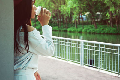 Side view of young woman having drink by railing against trees