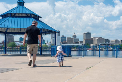 Rear view of father and daughter walking on land against sky in city