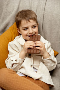 Little adorable boy sitting on the couch at home and eating chocolate bar. child and sweets, sugar