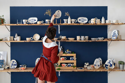 Back view of ceramist girl tidies up the shelves with handcraft ceramics at her studio