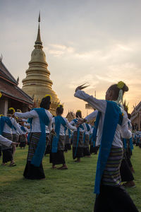 Women performing traditional dance by wat phra against sky during sunset