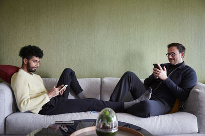 Gay couple relaxing on sofa at home and using phones