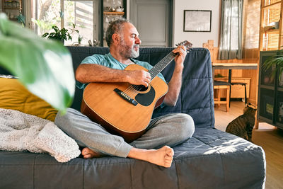 Middle-aged bearded man playing acoustic guitar while sitting on sofa in light living room.