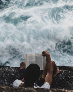 Directly above shot of man reading book while sitting on cliff against sea