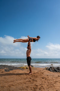 Full body shirtless man standing on sandy beach near waving sea and holding barefoot girlfriend in raised arms against cloudy blue sky on sunny summer day