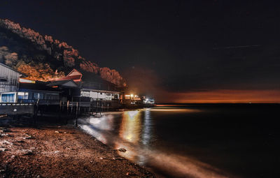 Illuminated buildings by sea against sky at night