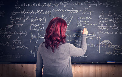 Rear view of teacher writing on blackboard in classroom