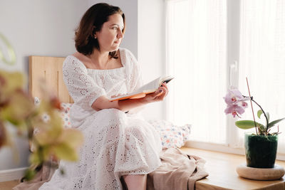 Mid age adult woman plus size reading a book by the window on the windowsill in the morning