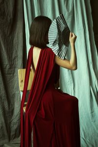 Fashion model holding hand fan while sitting on chair against curtains