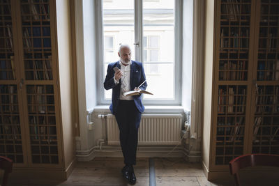 Senior lawyer reading book while listening through headphones against window in library
