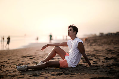 Full length of young woman sitting on sand at beach against sky