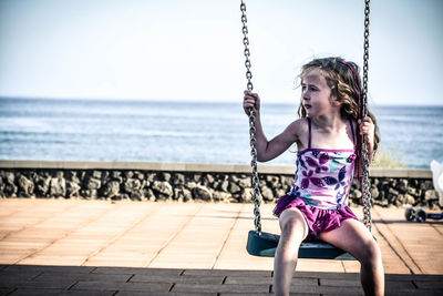 Cute girl looking away while sitting on swing against sea