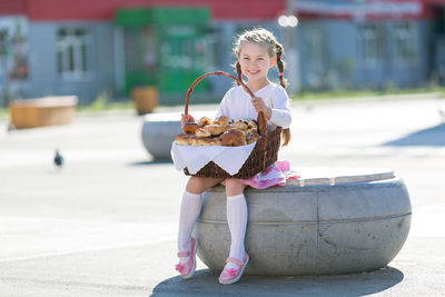 Full length of smiling girl holding basket with food while sitting on road