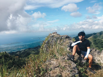 Young man sitting on mountain peak against cloudy sky