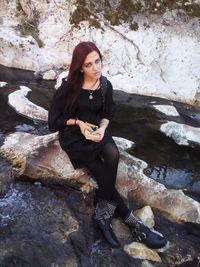 High angle portrait of young woman holding book while sitting on rock at beach