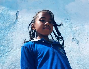 Low angle portrait of girl standing against blue wall during sunny day