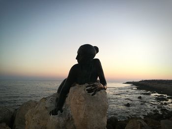 Silhouette person on rock by sea against clear sky during sunset