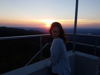 Woman standing by railing against sky during sunset