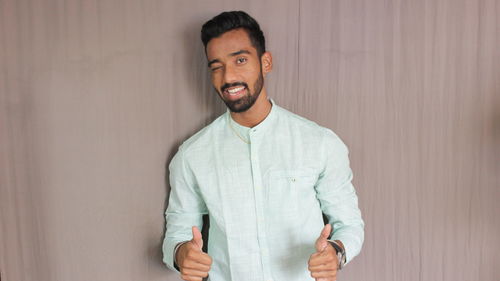 Portrait of young man showing thumbs up while standing against wall