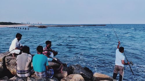 Rear view of people sitting on rocks by sea against clear sky