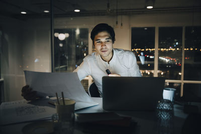 Confident businessman holding paper while looking at laptop in office
