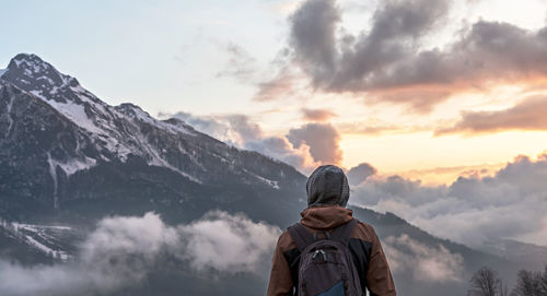 Young man traveler in hood with backpack from behind looking mountain view of the snow capped peaks