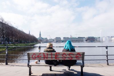 Rear view of women sitting on bench at alster river against sky