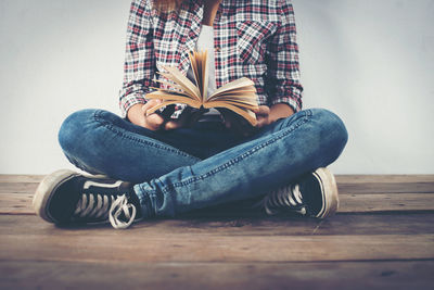 Low section of young woman holding book while sitting against wall on wooden floor